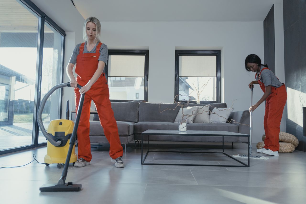 services-02 Dvě ženy v červených overalech vysávají a zametají moderní obývací pokoj.